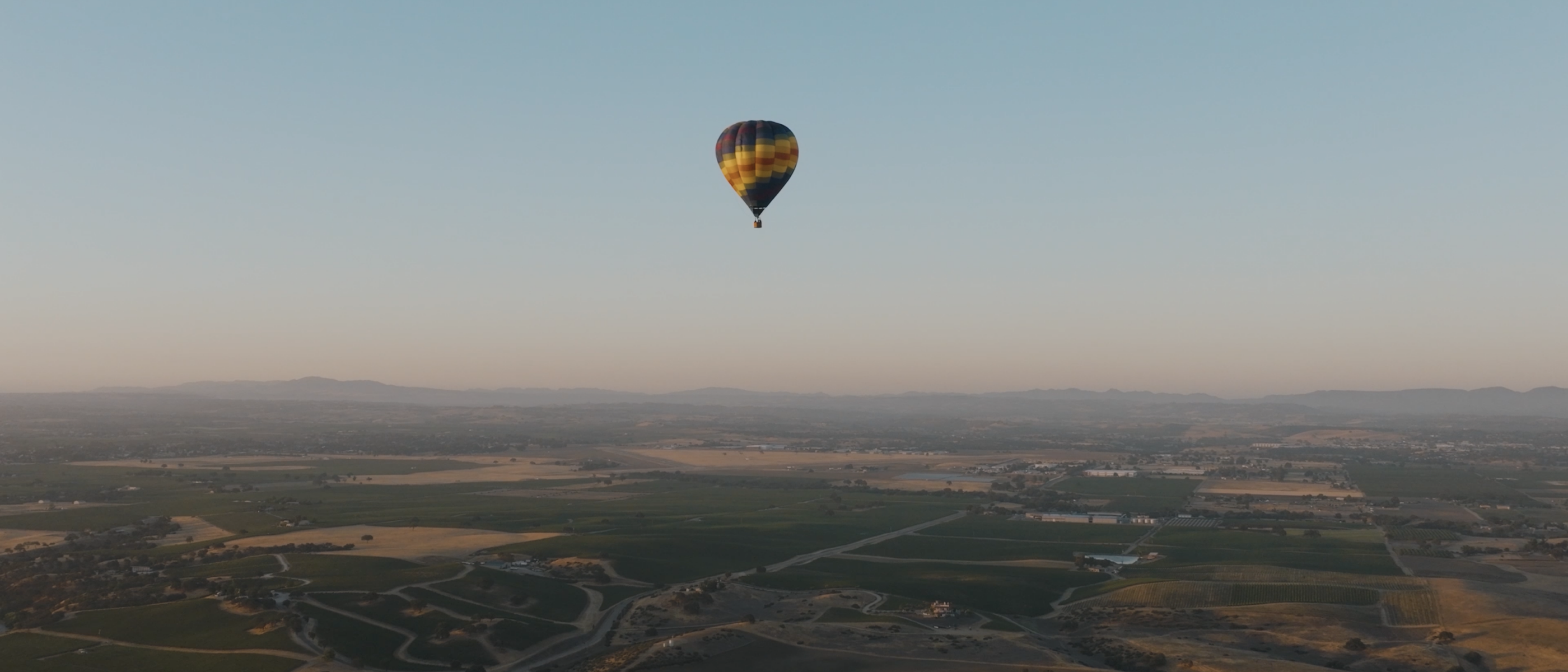 A colorful hot air balloon floats above a wide landscape of fields and hills under a clear sky.
