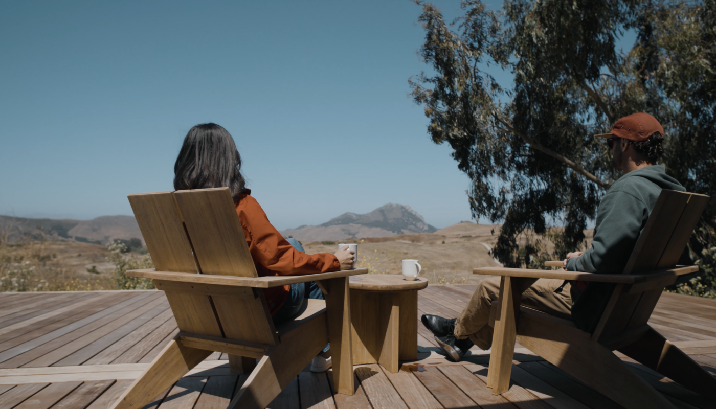Two people sit in wooden chairs on a deck, enjoying a scenic view of hills and mountains under a clear blue sky.