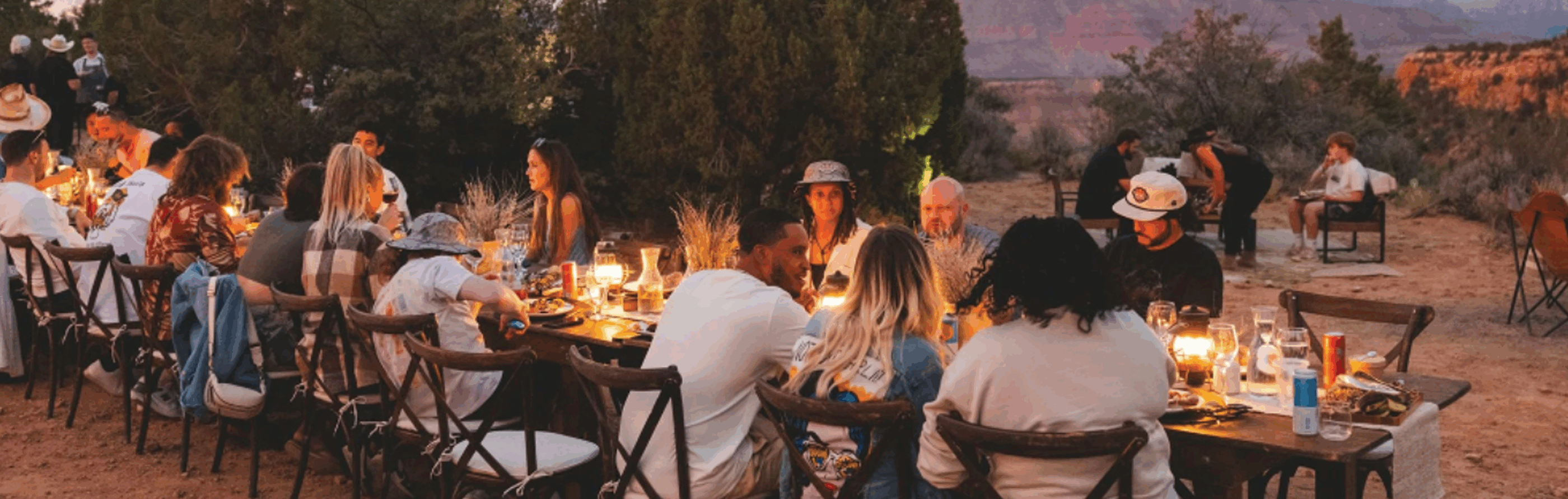 A long dining table is set outdoors in a desert landscape with guests enjoying a meal, surrounded by trees and mountains at sunset.