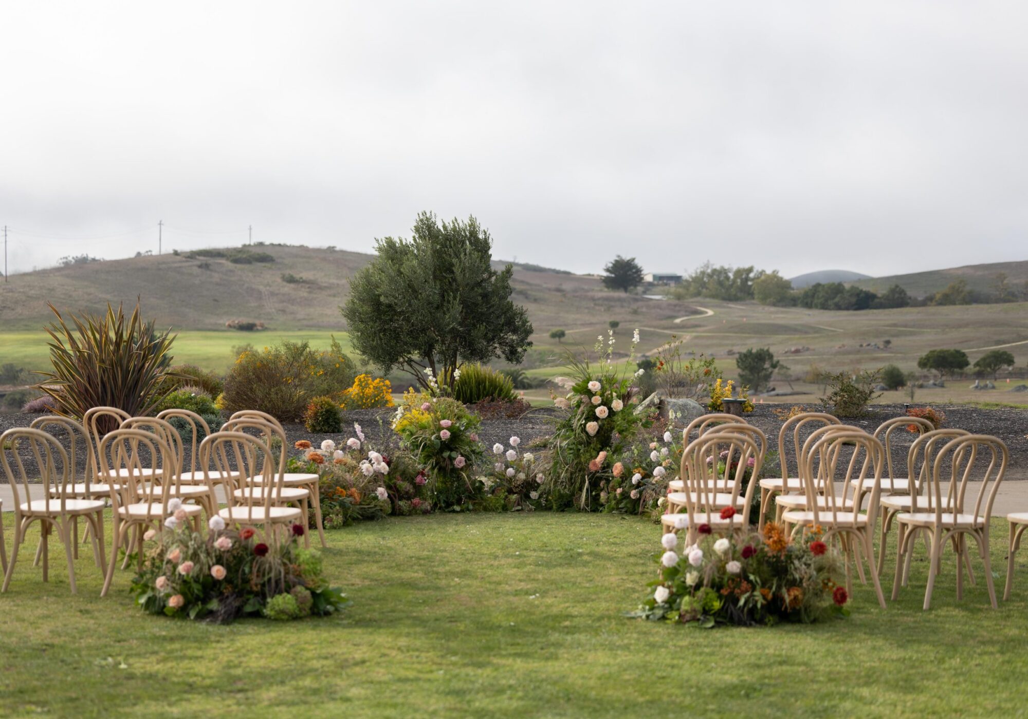 A circular arrangement of chairs surrounded by colorful flowers on a lawn, backed by a scenic hilly landscape under cloudy skies.