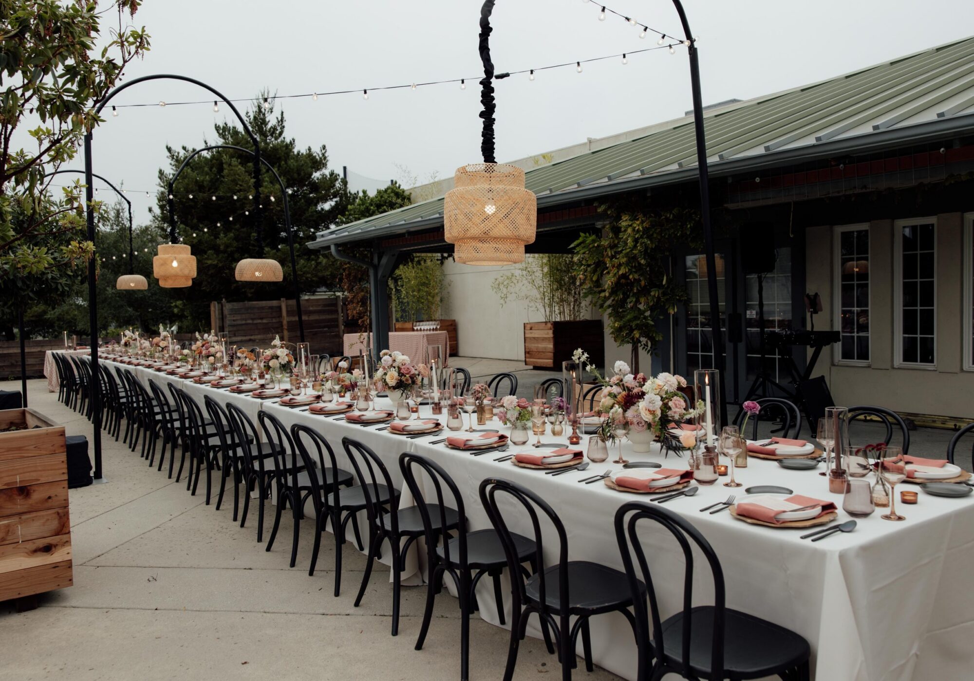 A long outdoor dining table is set for a gathering, adorned with flowers and tableware, under hanging lights.