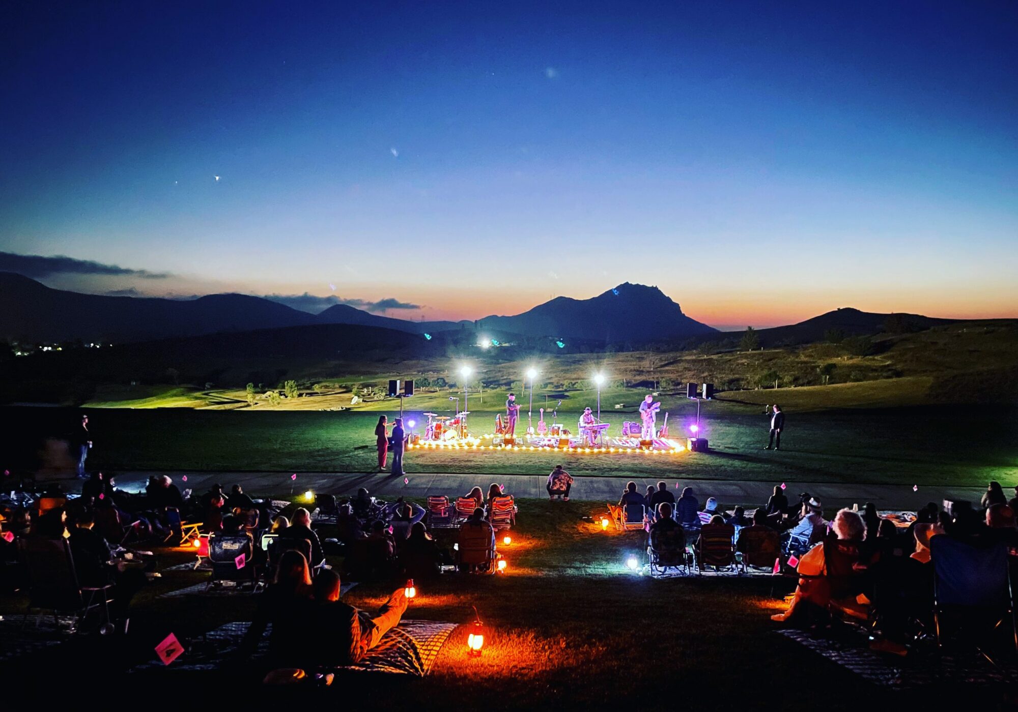 A crowd sits on a grassy hillside, watching a performance on stage illuminated by colorful lights at dusk, with mountains in the background.