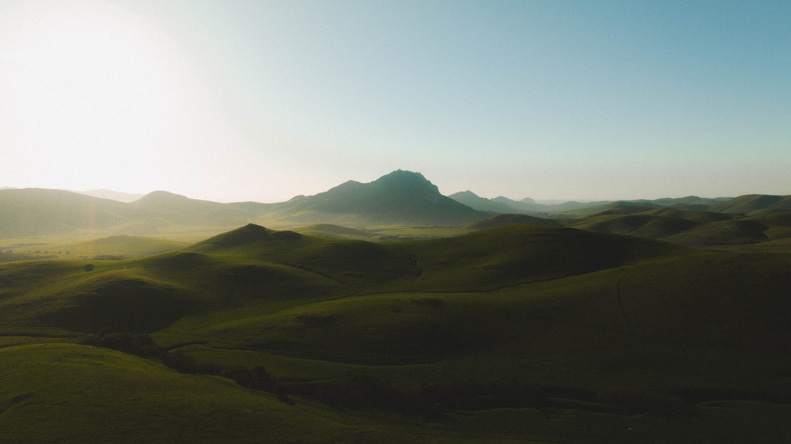 Lush green hills stretch across the landscape under a clear sky, with a distant mountain rising in the background.