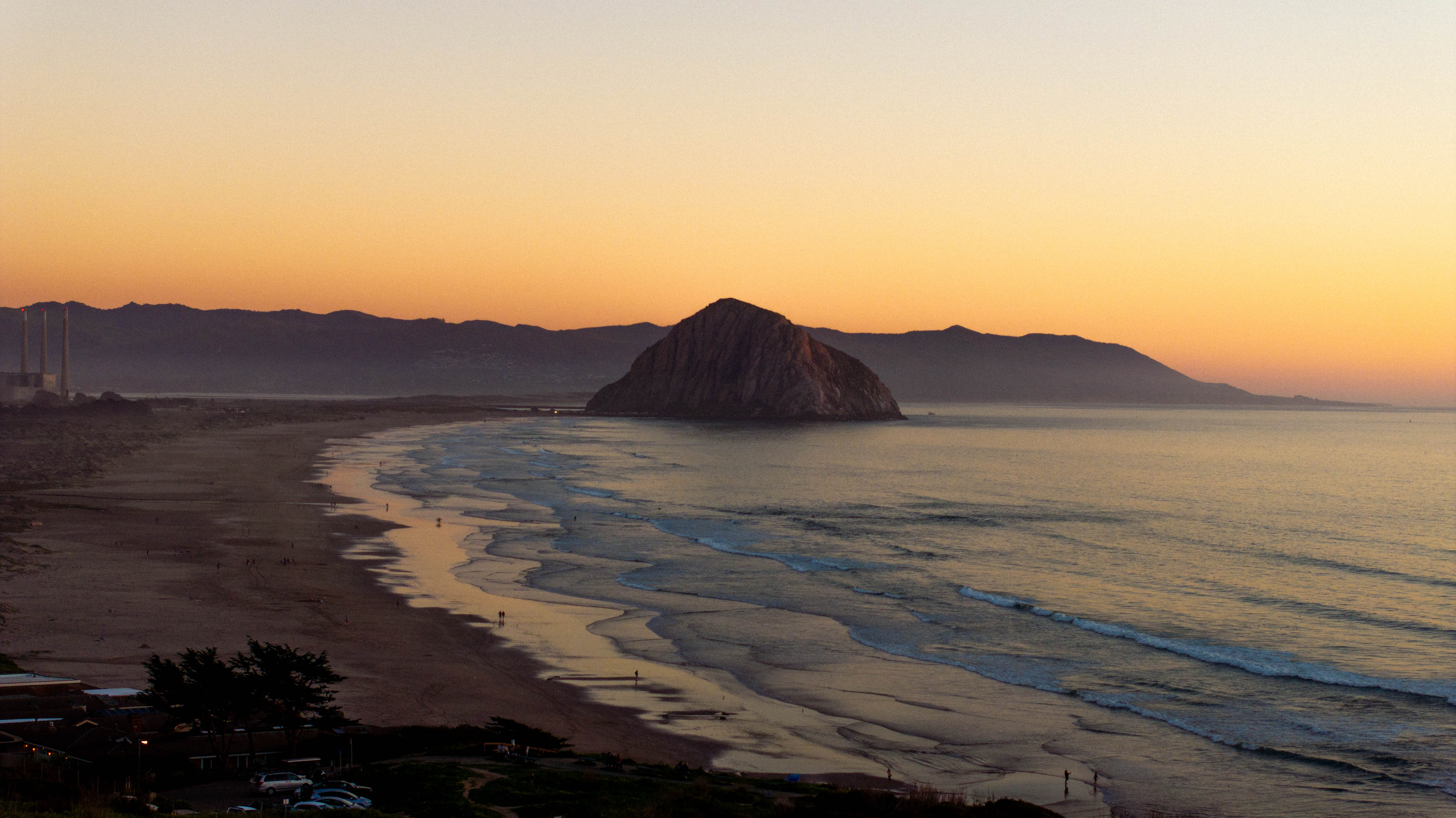 Sunset over a beach with gentle waves, featuring a prominent rock formation in the water and distant mountains.