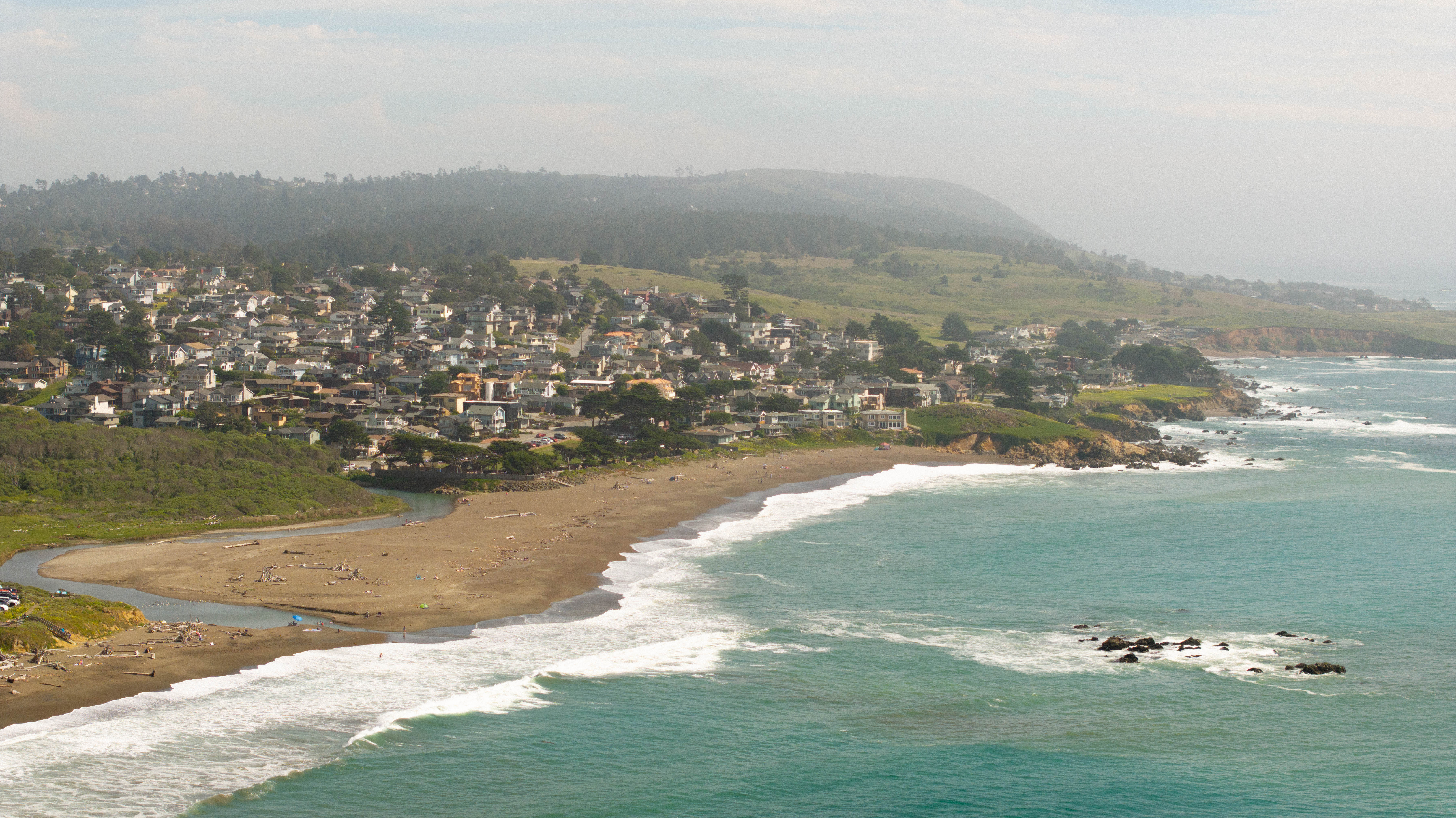 Coastal view of a small town by the ocean, with rolling hills and waves lapping at the shoreline.