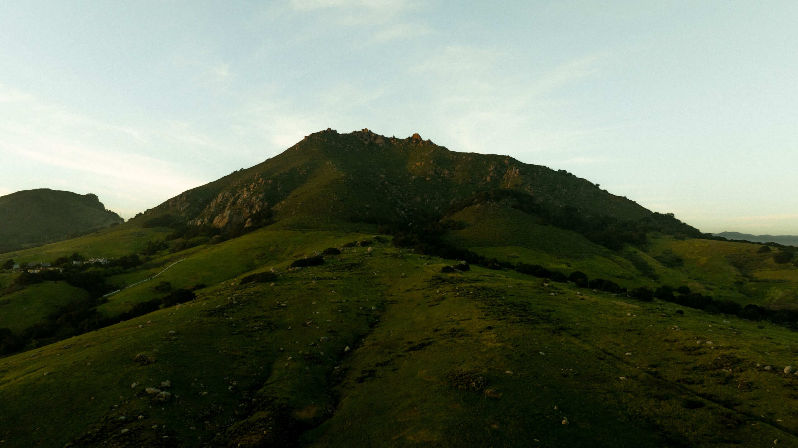 Rolling green hills lead up to a prominent rocky peak under a clear sky. The landscape appears tranquil and expansive.