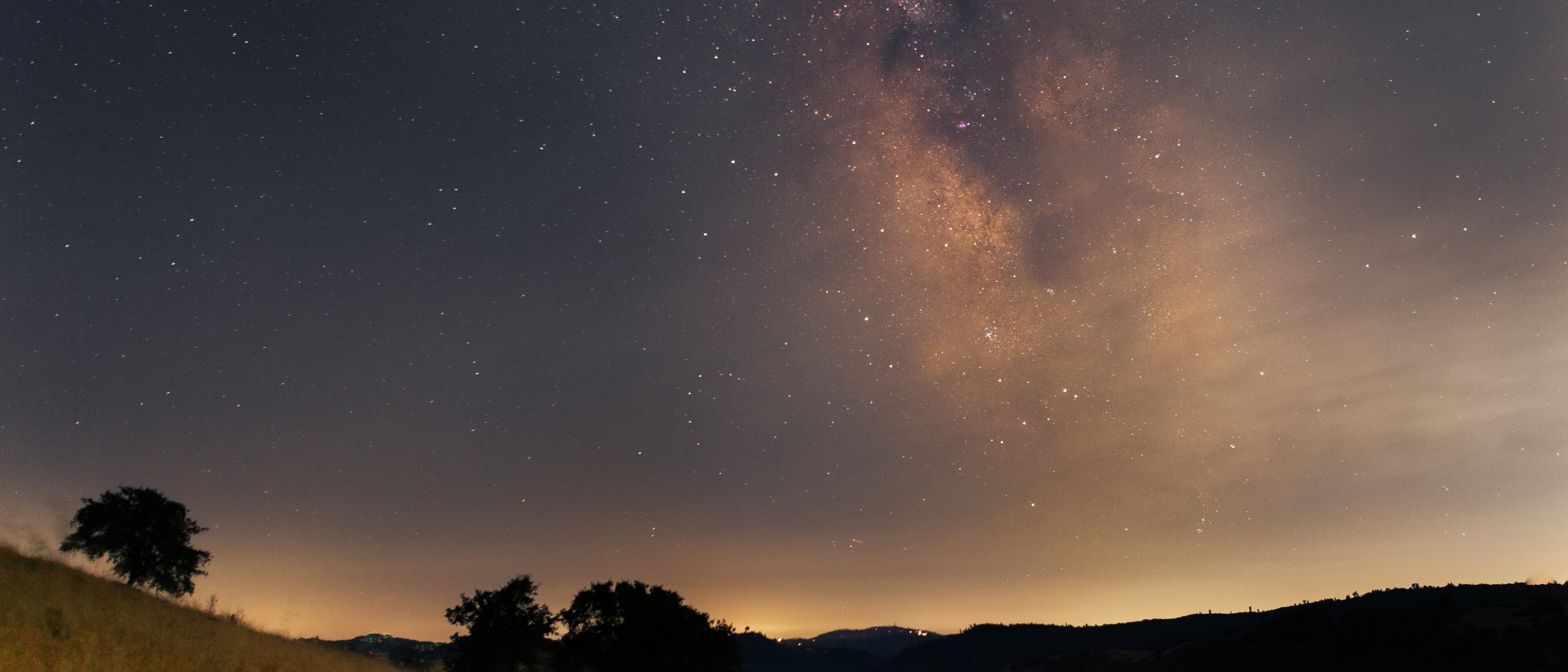 The Milky Way rising above golden grass hills and oak trees in California