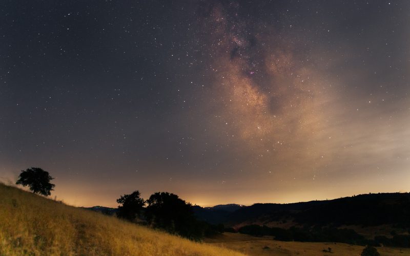The Milky Way rising above golden grass hills and oak trees in California